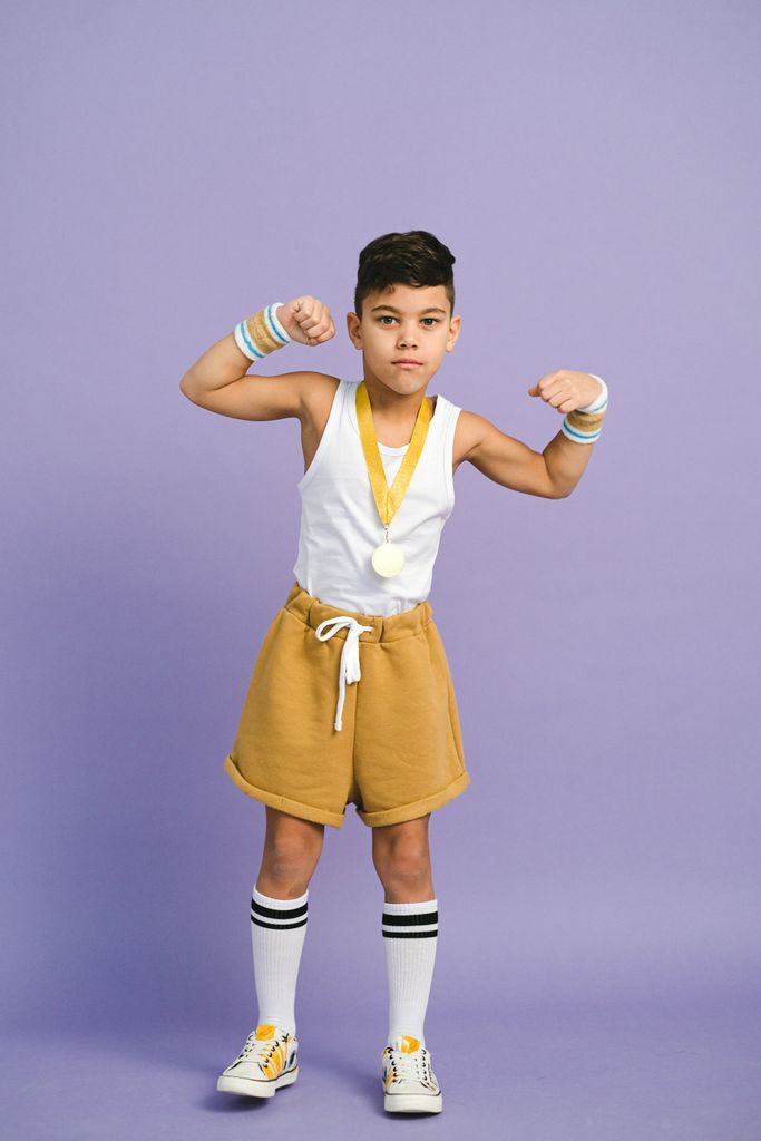 Confident young boy flexing his muscles wearing a gold medal, standing on a pink background.